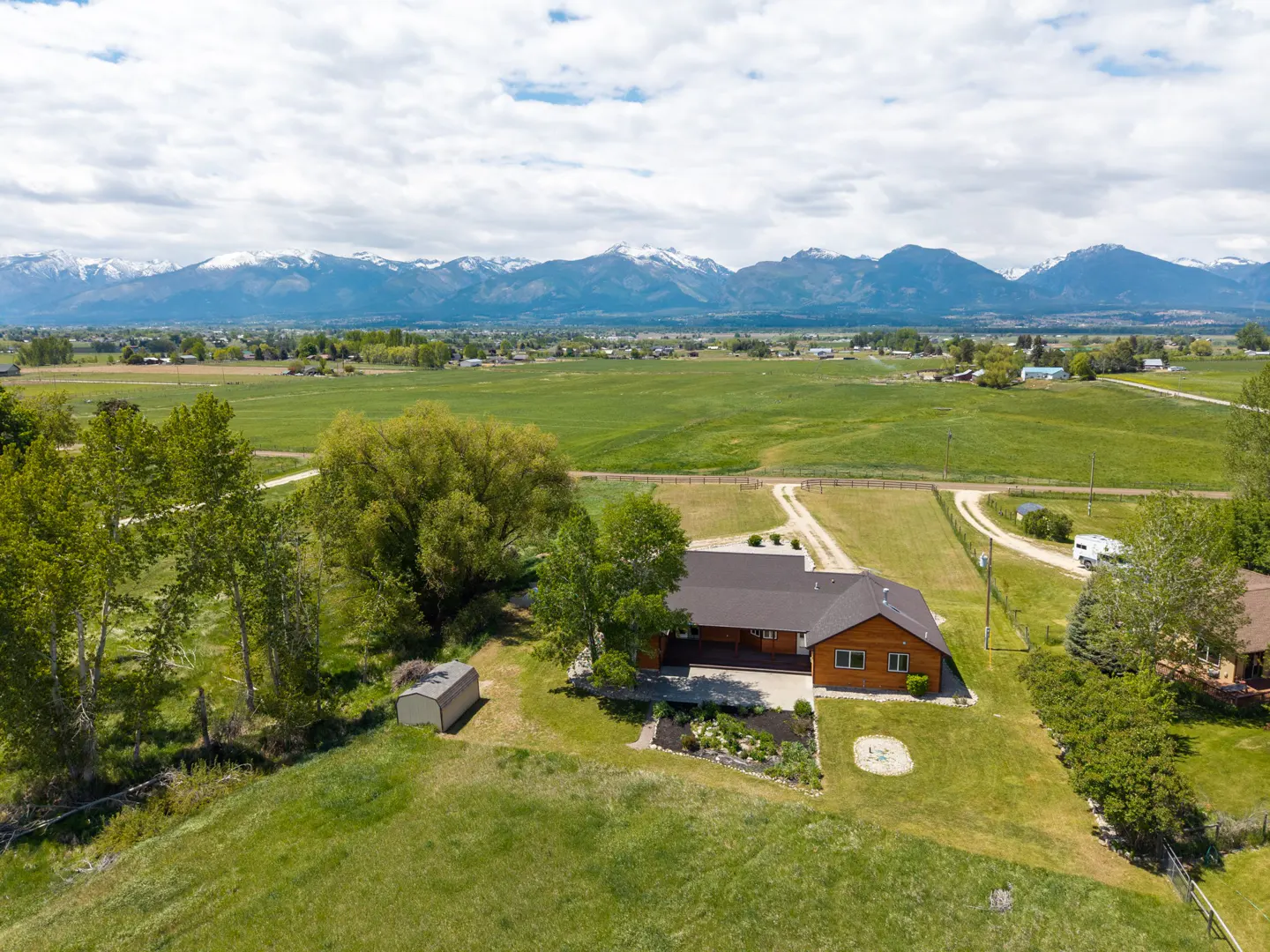 Aerial view of a brown ranch-style home on a large green lot with a shed, surrounded by fields and snow-capped mountains.