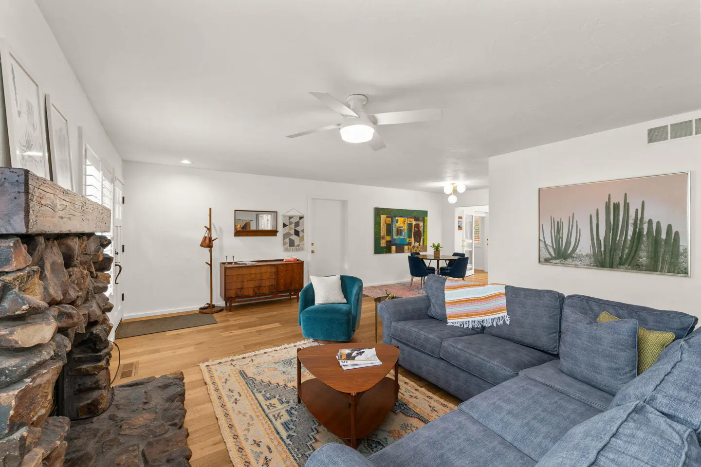 Bright living room with a stone fireplace, blue sectional sofa, wood floors, and white walls. A dining area is visible in the background.