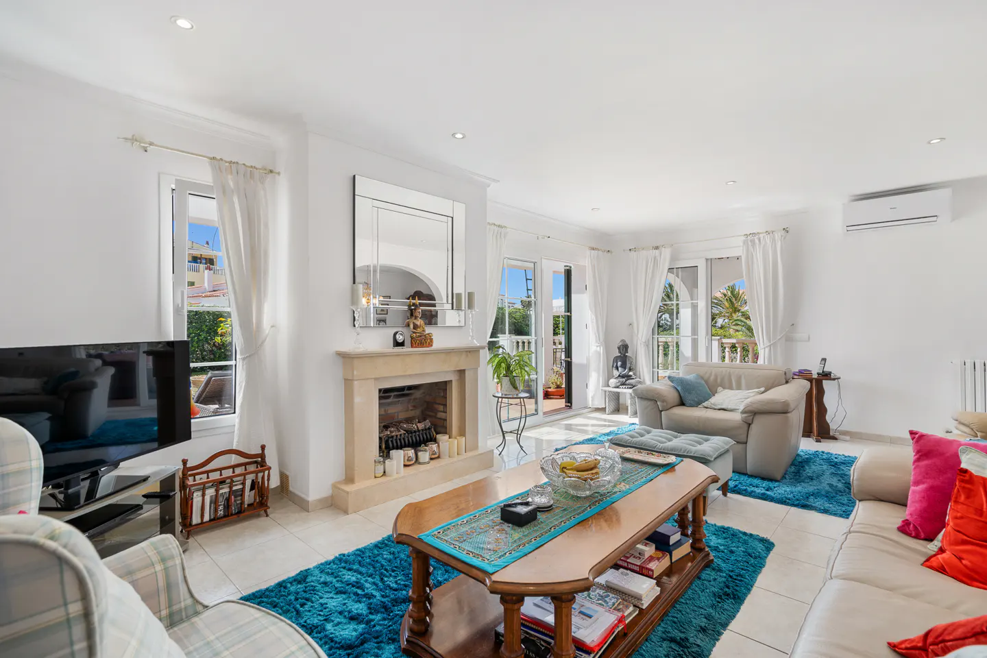 Bright living room with white walls, tile floors, and blue rugs. A fireplace, sofas, and a wooden coffee table furnish the space. Natural light streams through windows.