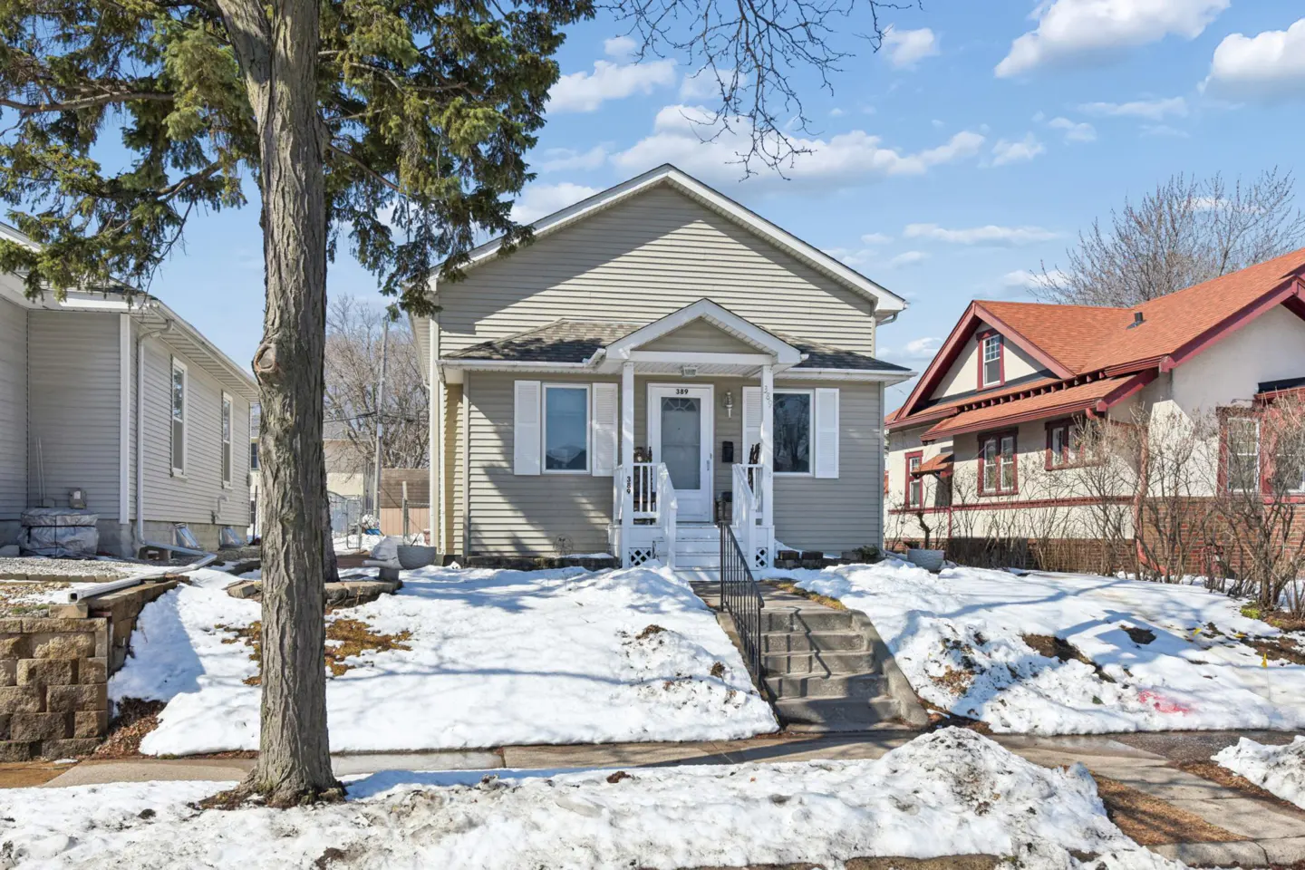 A beige, two-story house with white trim and a small porch, sits on a snowy lawn under a blue sky.