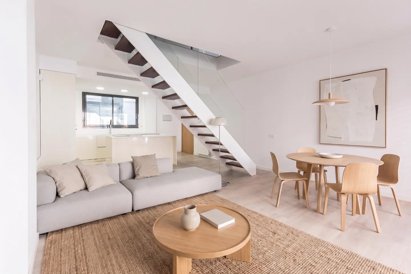 Bright, modern living room with a gray sofa, round wood table, and a dining set. A white staircase with glass railing leads upstairs.