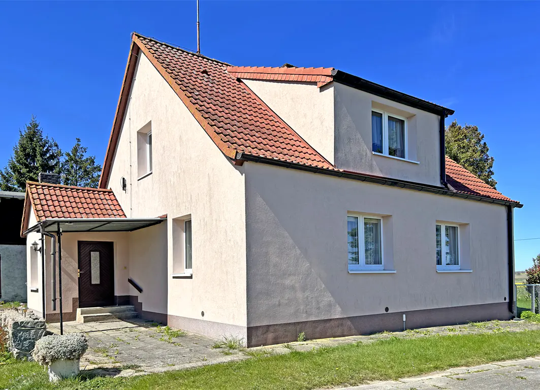 Two-story house with a red tile roof and a beige exterior under a clear blue sky. A covered porch leads to the front door.