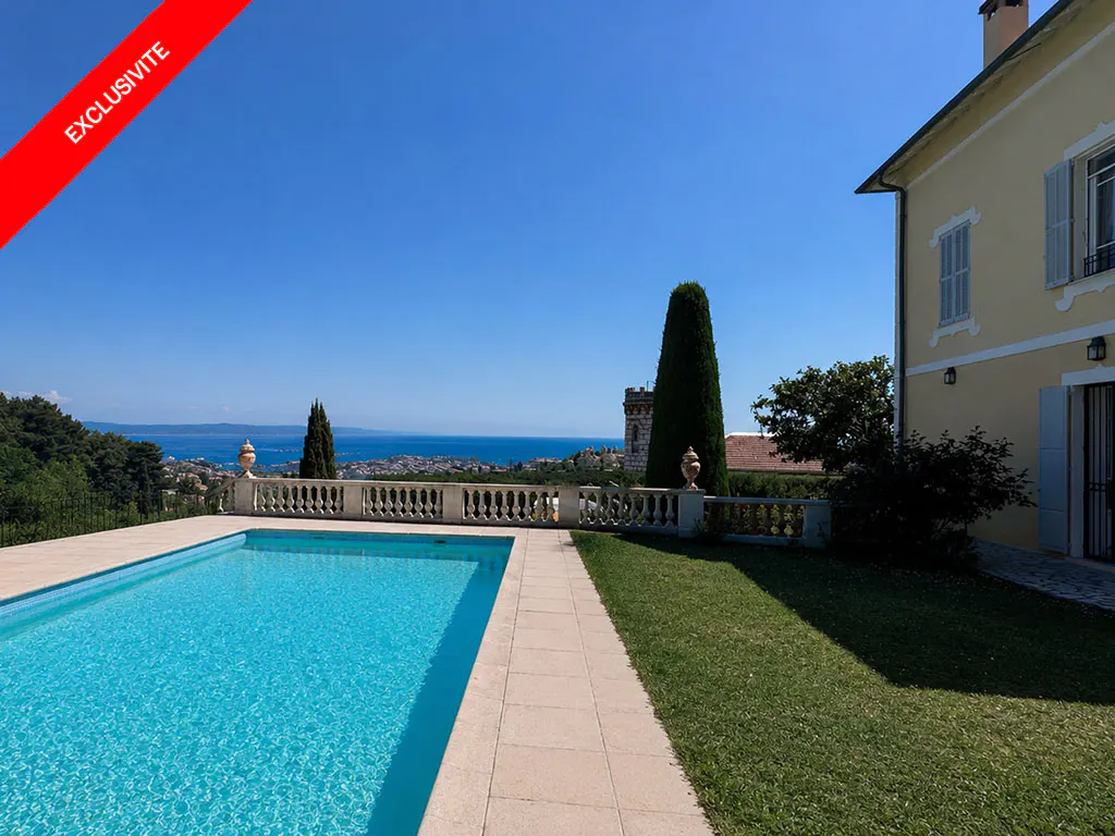 A sunny view of a pool, lawn, and yellow house overlooking the sea. A red "Exclusive" banner is in the upper left corner.