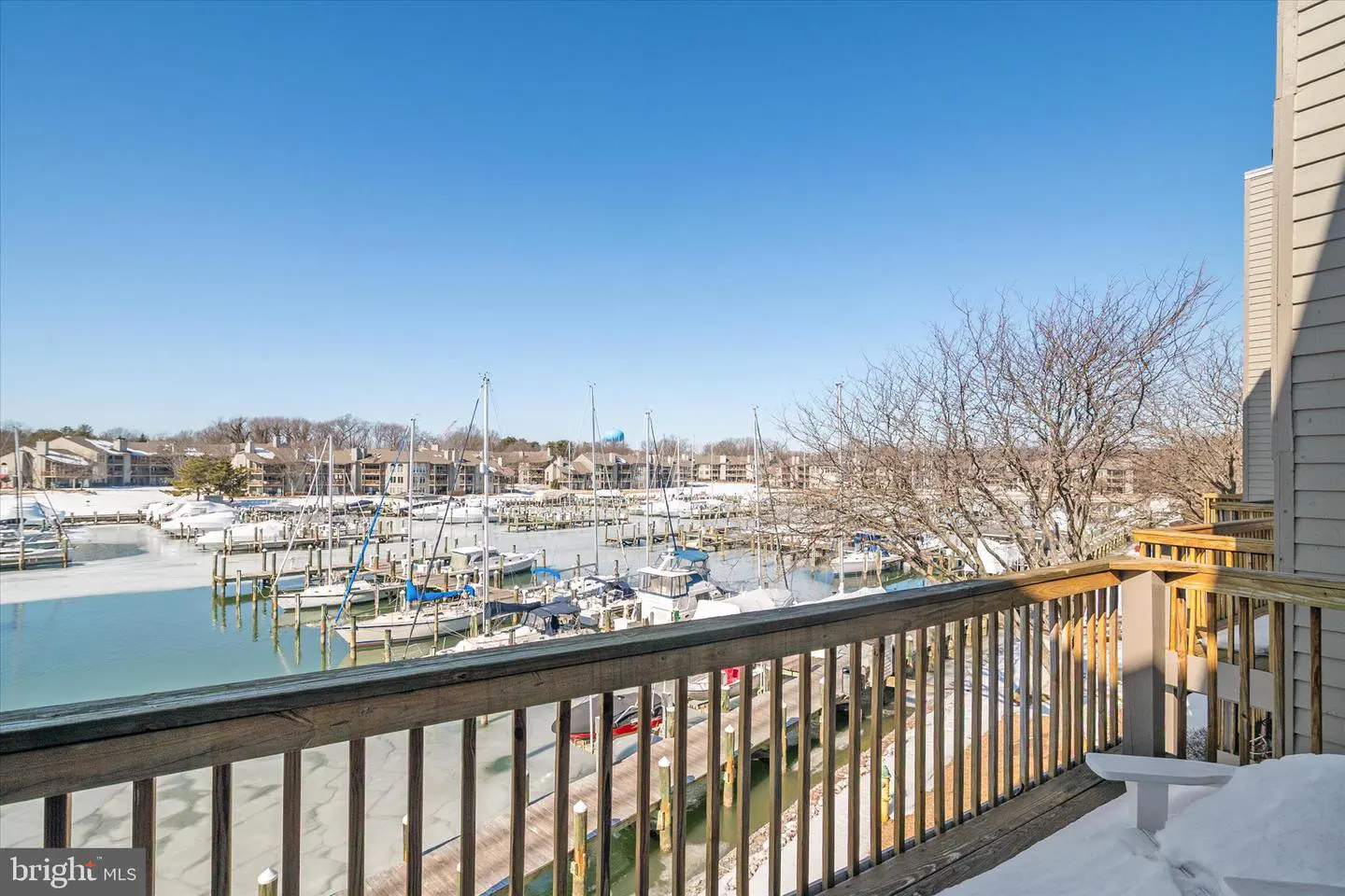 View from a wooden balcony overlooking a marina with snow-covered boats and docks under a clear blue sky.