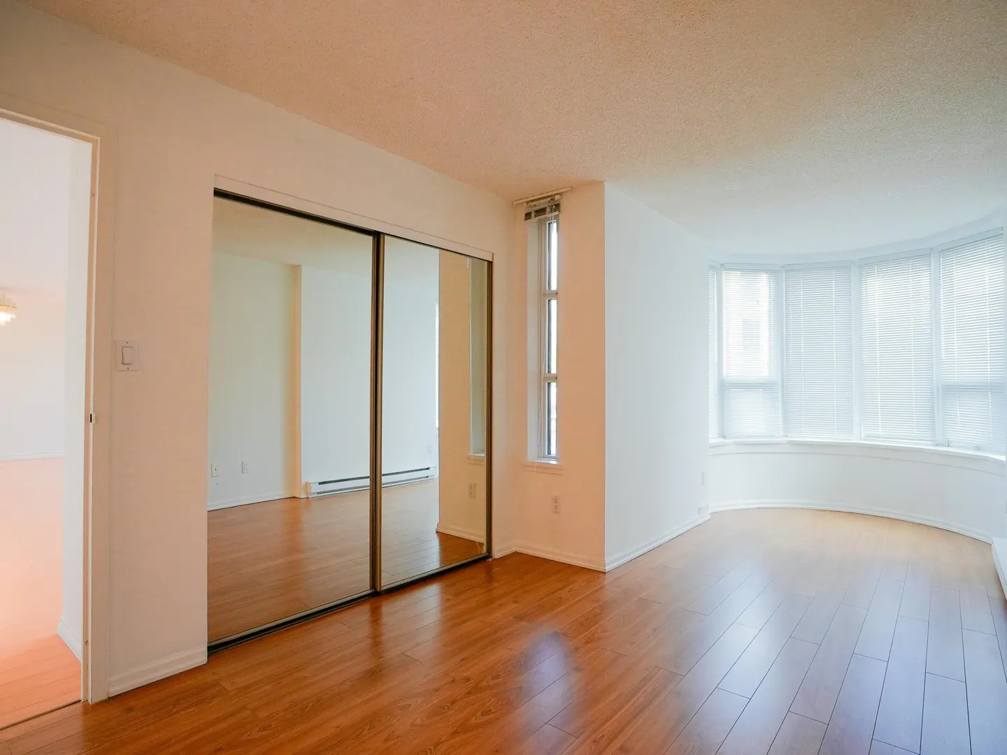 Bright, empty room with wood floors, white walls, and a bay window. Mirrored closet doors reflect the adjacent room.