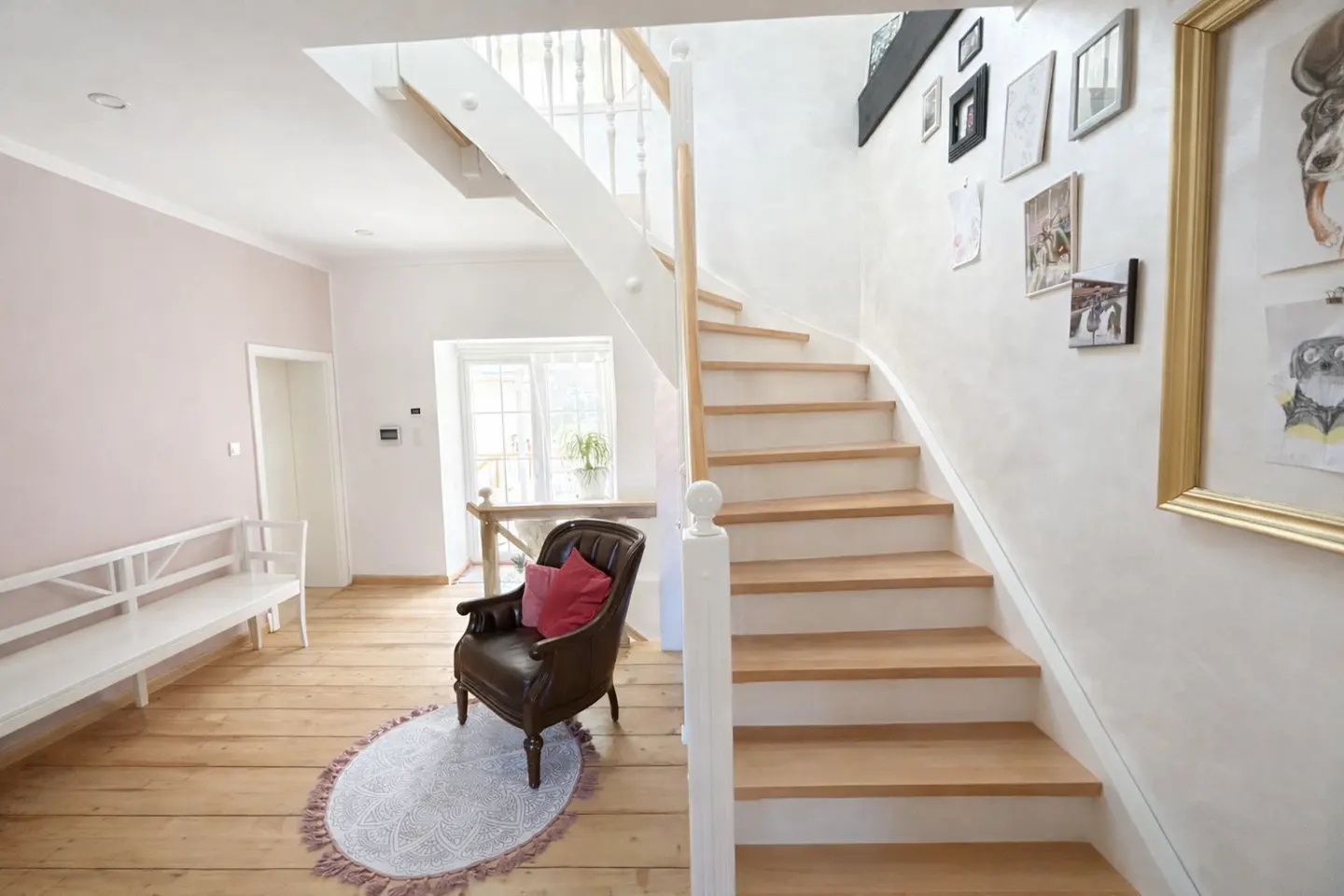 Bright foyer with wooden floors, white walls, and a staircase. A brown leather chair with a red pillow sits on a round rug. Artwork adorns the walls.