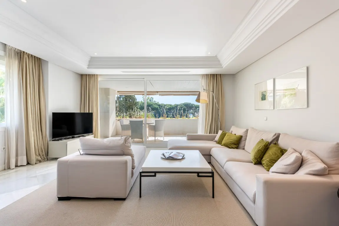 Bright living room with a white sectional sofa, green accent pillows, a coffee table, and a view of a balcony with trees.