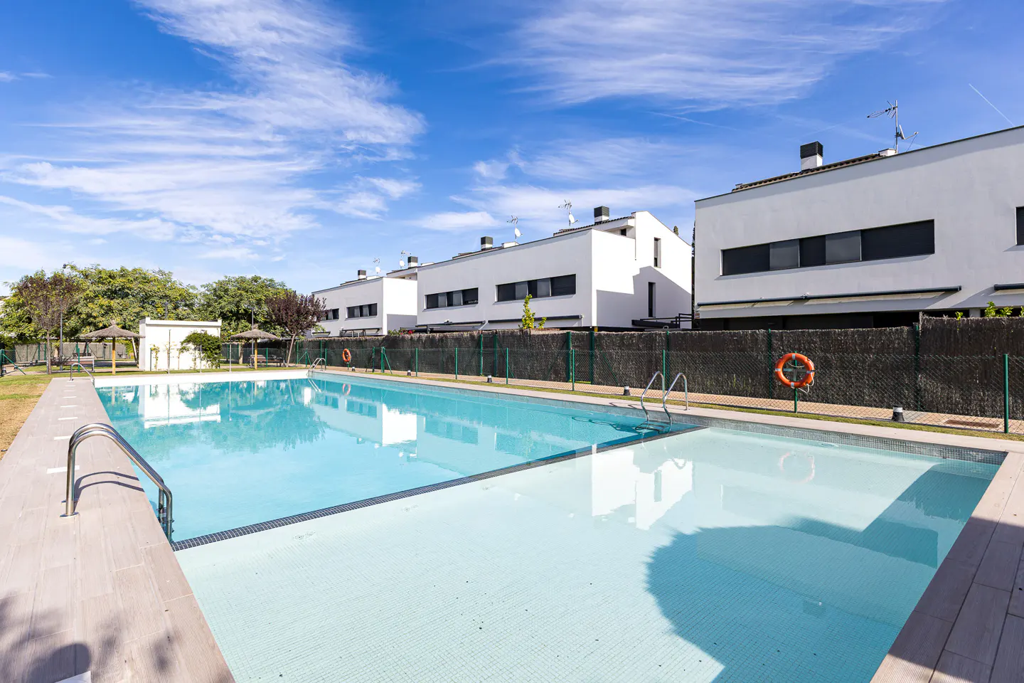A bright, sunny day over a community pool. White modern townhouses line the background behind a dark fence. A life preserver hangs on the fence.