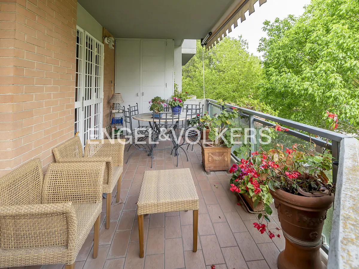 A balcony with wicker chairs, a table, and potted red flowers. Green trees are visible in the background.