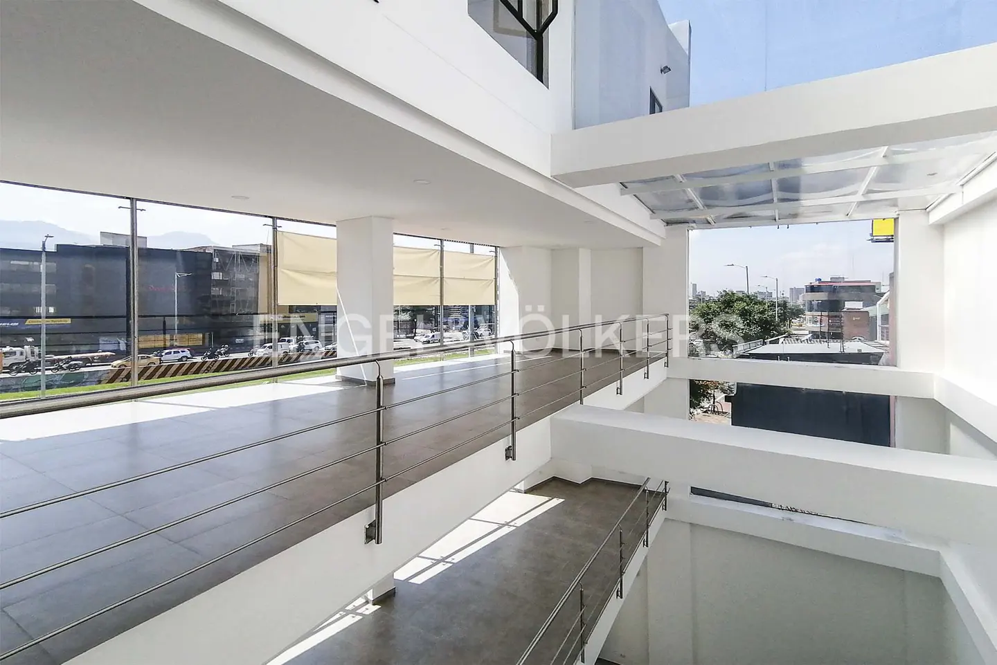 Modern building interior with white walls, gray floors, and metal railings. Large windows offer city views.