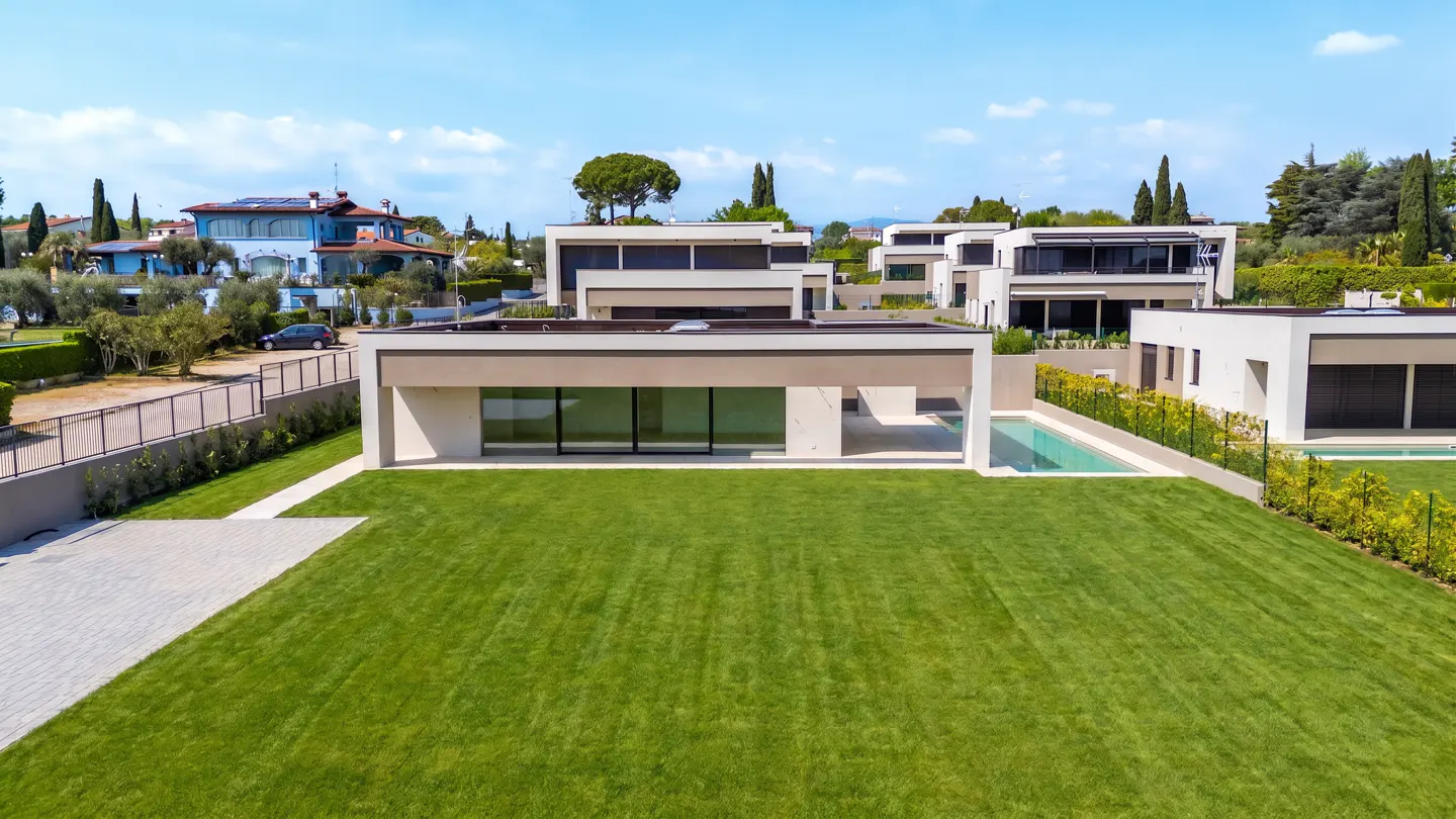 Modern white homes with flat roofs and large windows overlook a green lawn and swimming pools under a blue sky.