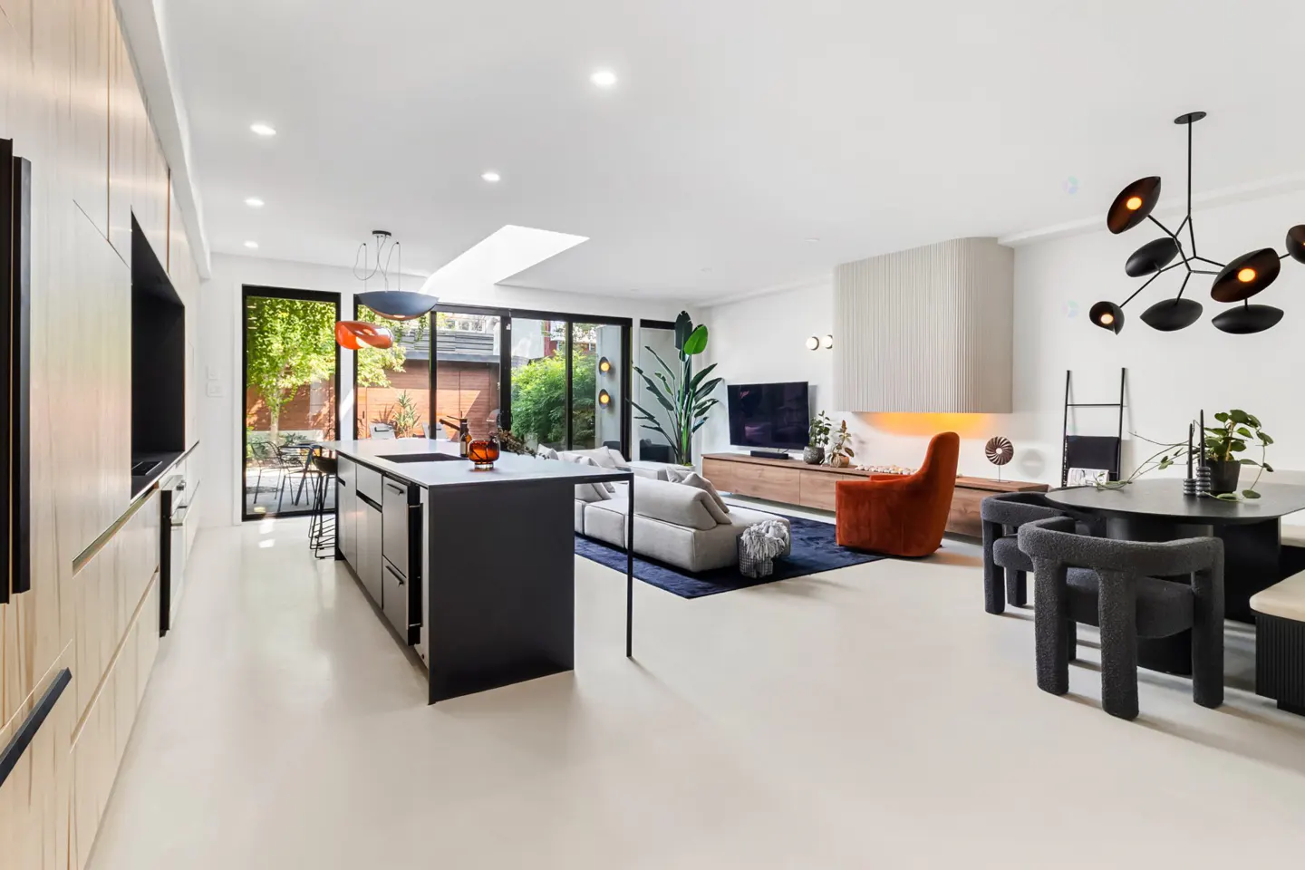 Bright, modern open-concept living space with a black kitchen island, white sofa, orange chair, and black dining table. Large windows offer a view of the backyard.