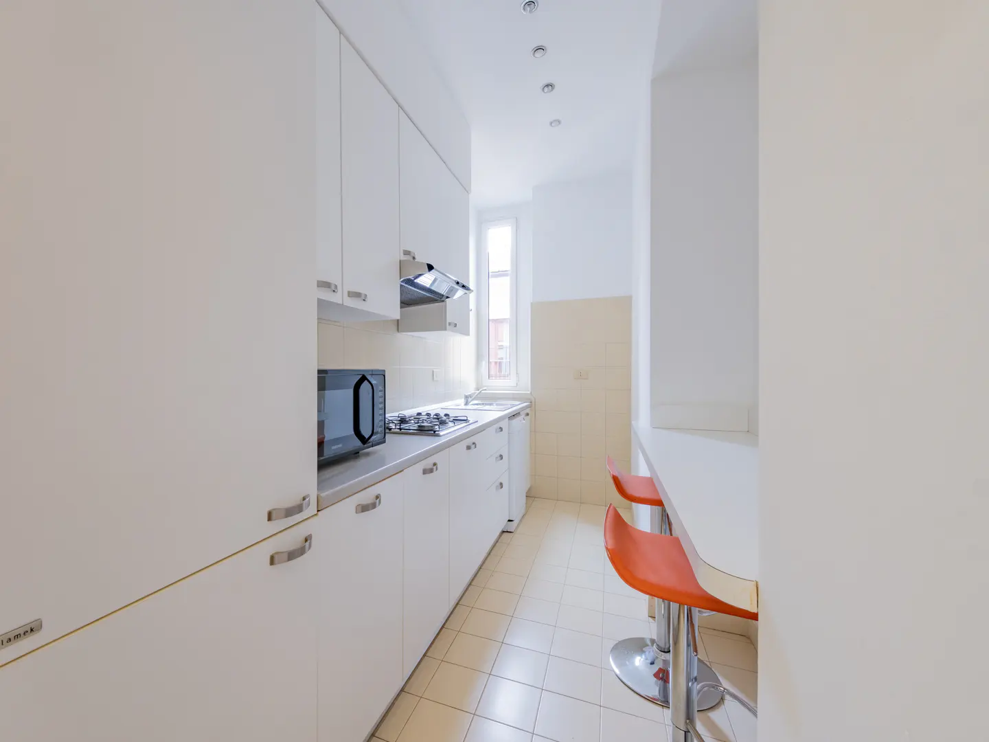 A bright, narrow kitchen with white cabinets, appliances, and tile floor. Two orange bar stools sit at a white counter.