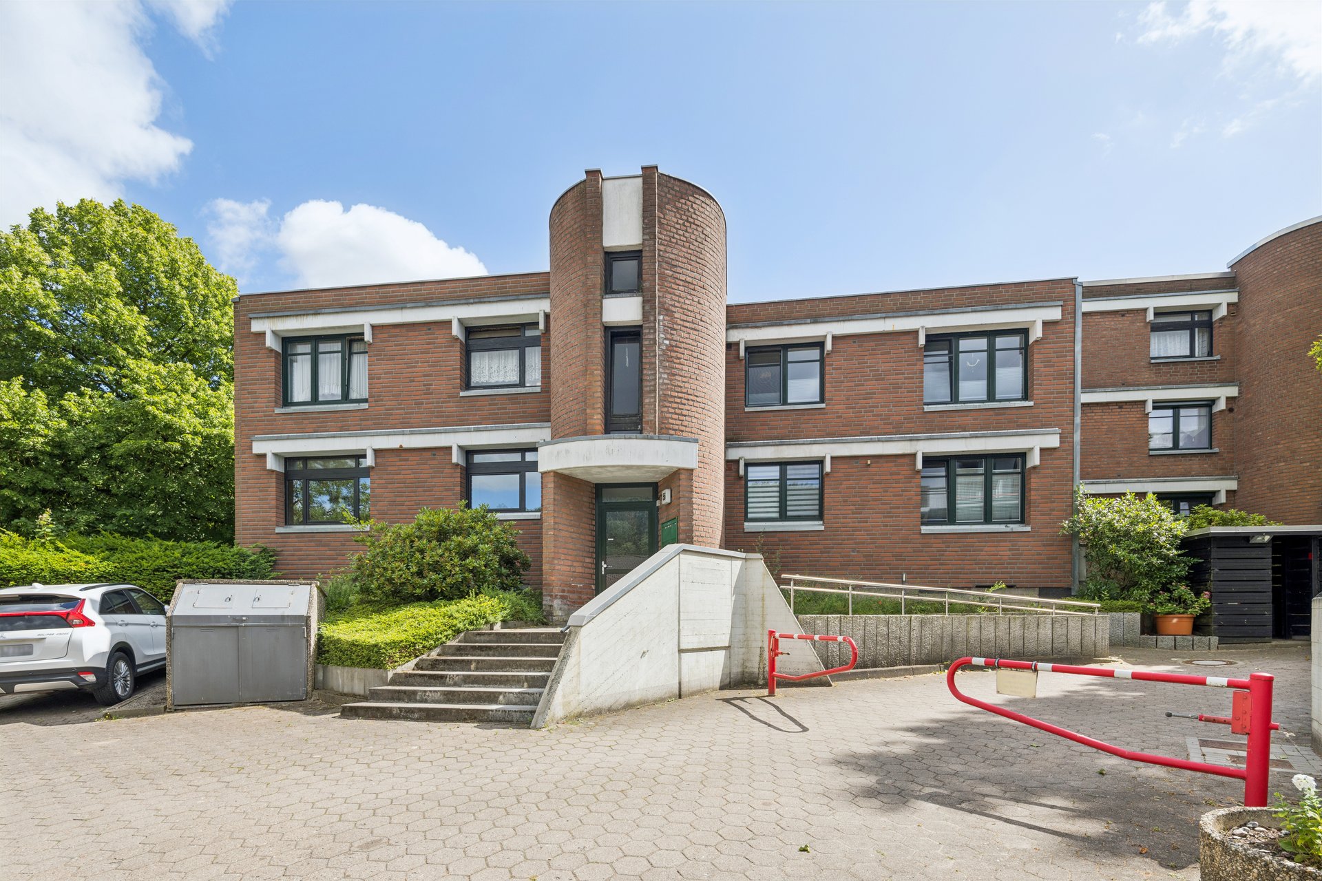 Exterior of a two-story red brick building with a central cylindrical tower, concrete stairs, and a red parking barrier.