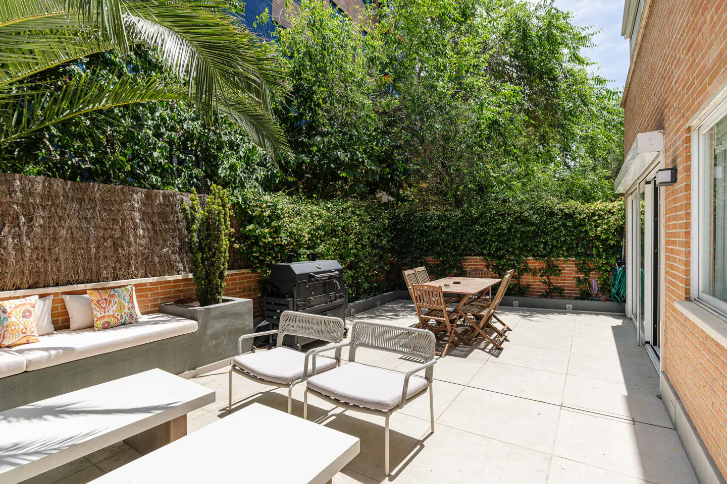 Sunny patio with a built-in bench, modern chairs, dining set, and grill against a brick wall and lush green hedge.