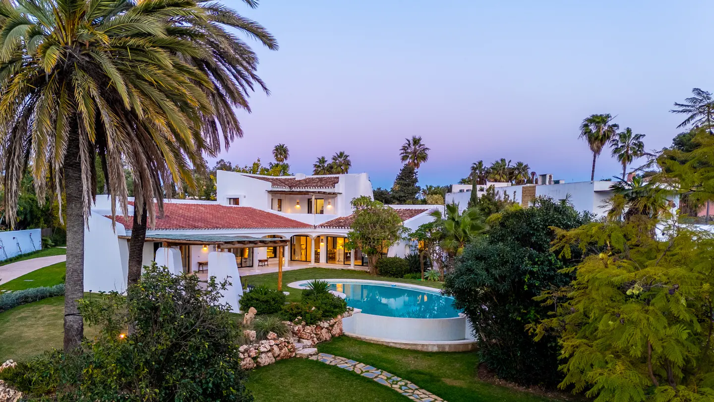 Exterior view of a white, Spanish-style home with a red tile roof, a pool, and palm trees at dusk.