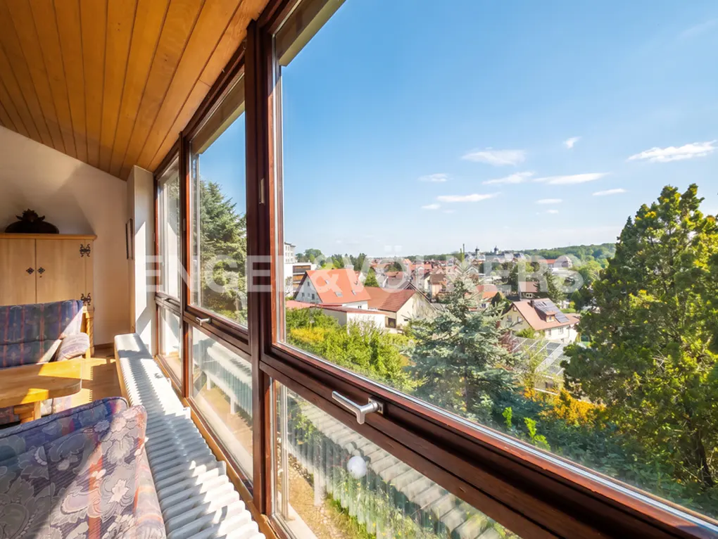 A bright room with a wood ceiling and a large window overlooking a town with trees and a blue sky.
