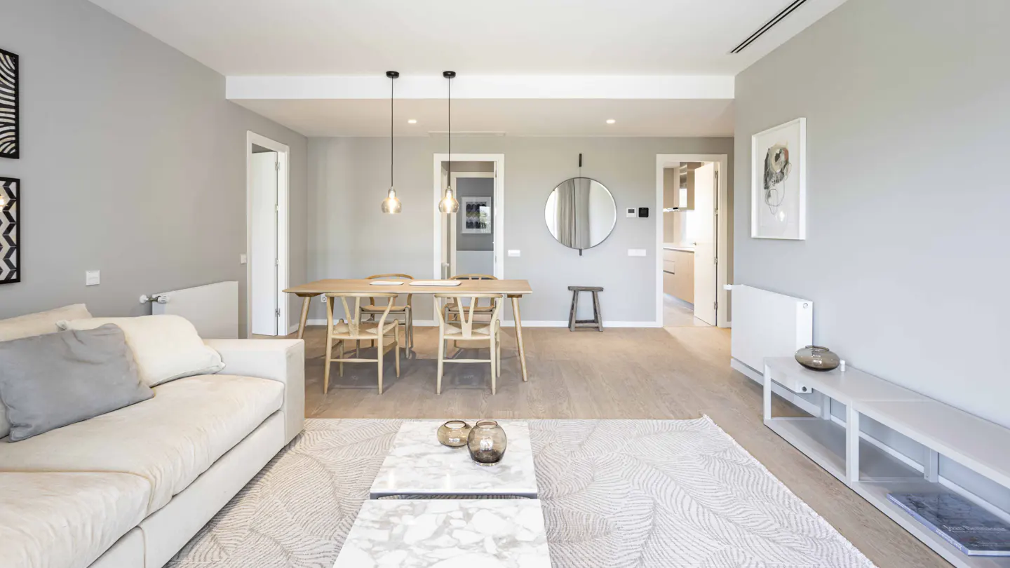 A bright, open-concept living and dining area with light gray walls, wood floors, and modern furniture. A white sofa, marble coffee table, and wooden dining set are visible.