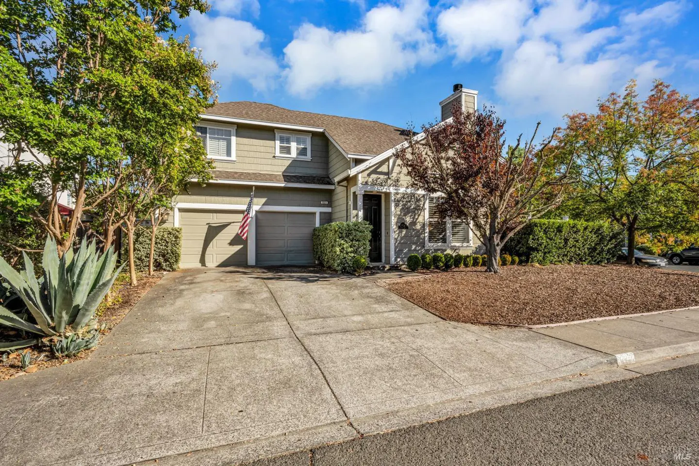 Two-story house with a gray exterior, a two-car garage, and an American flag. A tree with red leaves is in the front yard.
