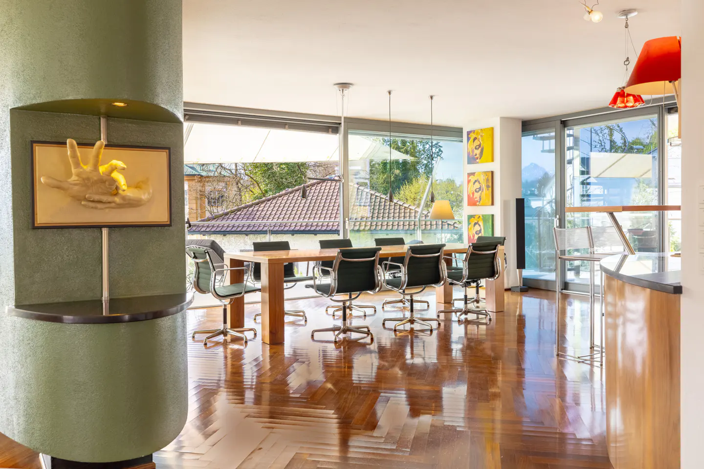Bright dining room with a long wooden table, green chairs, and a large window overlooking a cityscape. A green column with art is on the left.