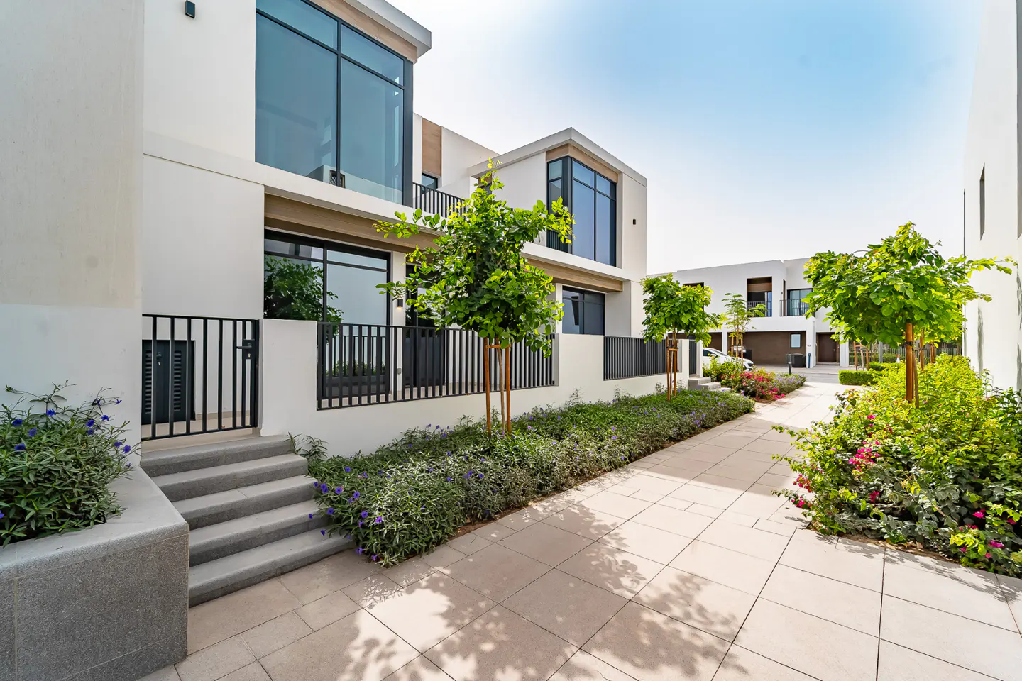 Modern white townhouses with black railings and large windows line a paved walkway with trees and flowers.