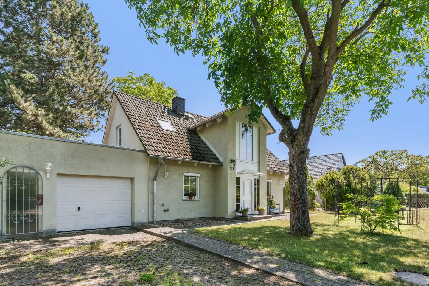 Two-story beige house with a brown tiled roof, white trim, and a two-car garage on a sunny day. A large tree is in the front yard.