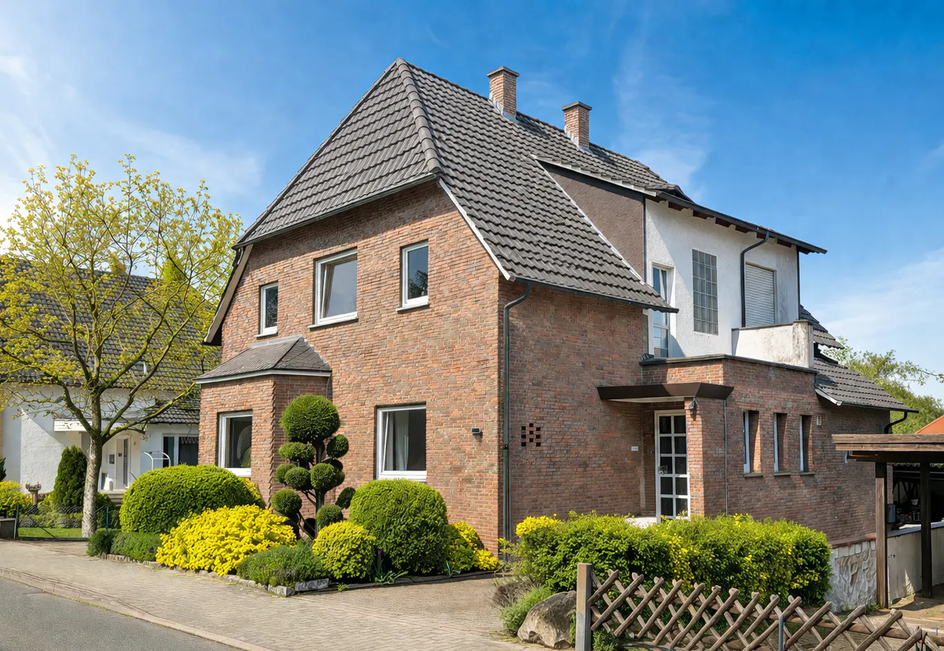 Two-story brick house with a gray roof, white windows, and manicured green and yellow bushes in the front yard under a blue sky.