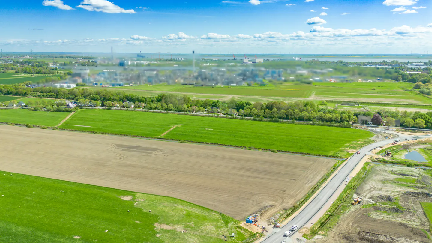 Aerial view of green and brown fields, a road under construction, and a blurred industrial area in the distance under a blue sky.