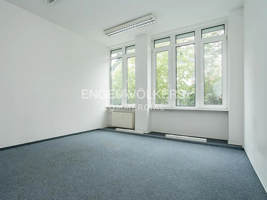 Empty office room with white walls, grey carpet, and large windows overlooking green trees.