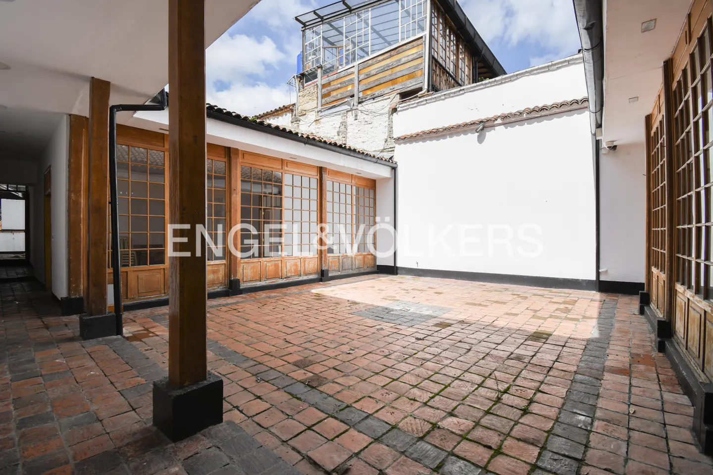 Outdoor courtyard with brick flooring, wooden pillars, and large windows. A white wall and a glass structure are visible in the background.