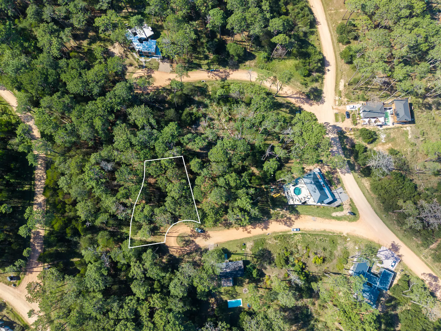 Aerial view of a wooded lot outlined in white, surrounded by green trees and dirt roads. Houses are visible in the background.