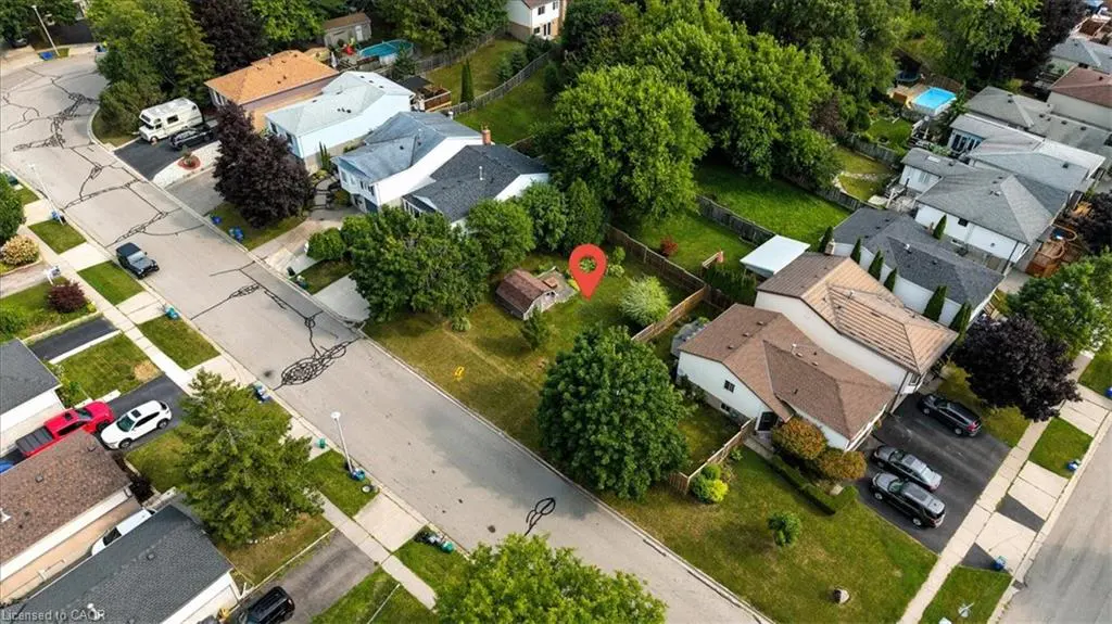 Aerial view of a residential street with houses, green trees, and lawns. A red pin marks a property with a small shed in the backyard.