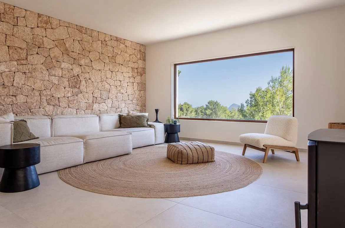 Living room with stone wall, white sofa, round jute rug, and large window with a view of trees.