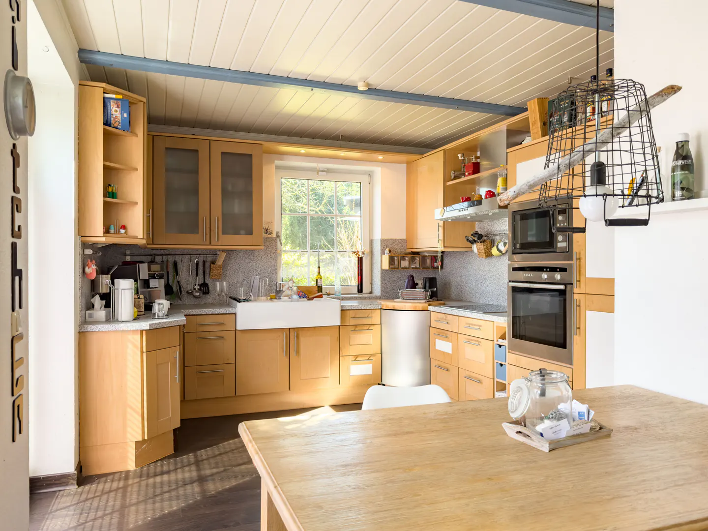 Bright kitchen with light wood cabinets, white farmhouse sink, and stainless steel appliances. A wooden table sits in the foreground.