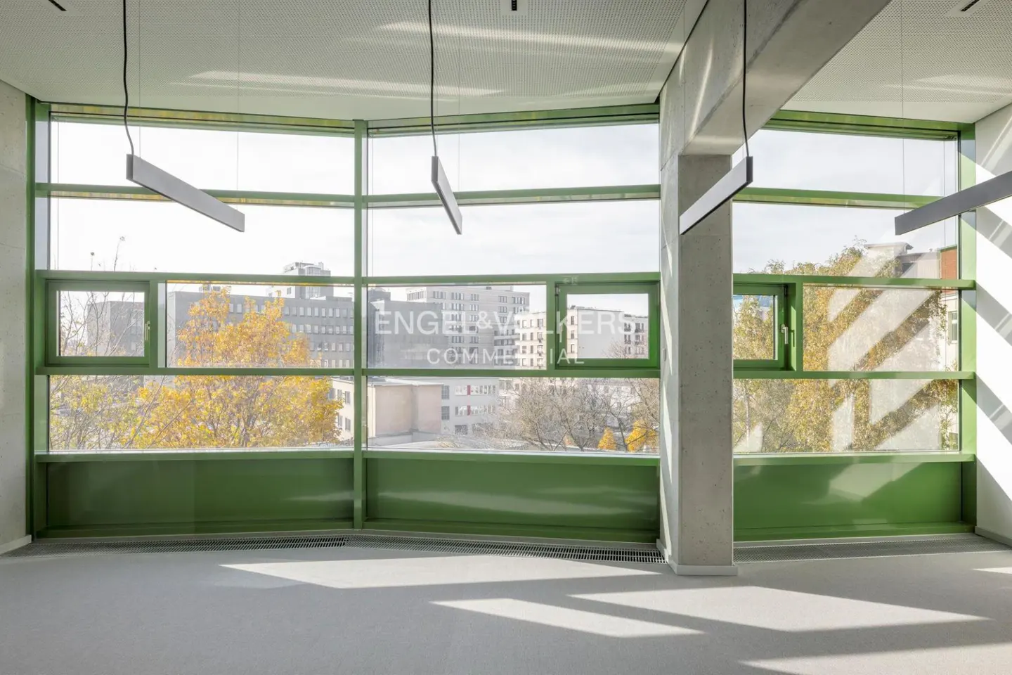 Empty room with green window frames, concrete pillars, and hanging lights. Outside, buildings and trees are visible.