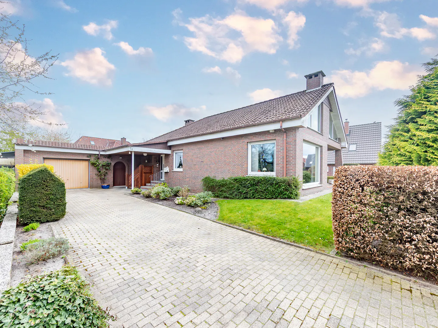 Exterior view of a one-story brick house with a paved driveway and manicured lawn under a blue sky with clouds.