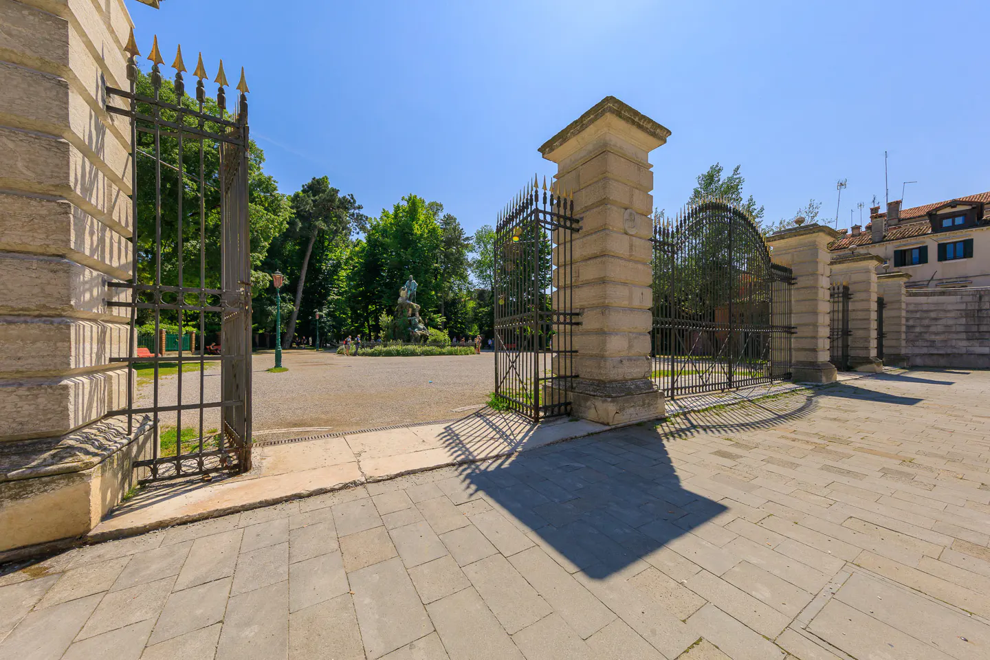 Open wrought iron gate with gold spikes, flanked by stone pillars, leads to a park with trees and a statue.