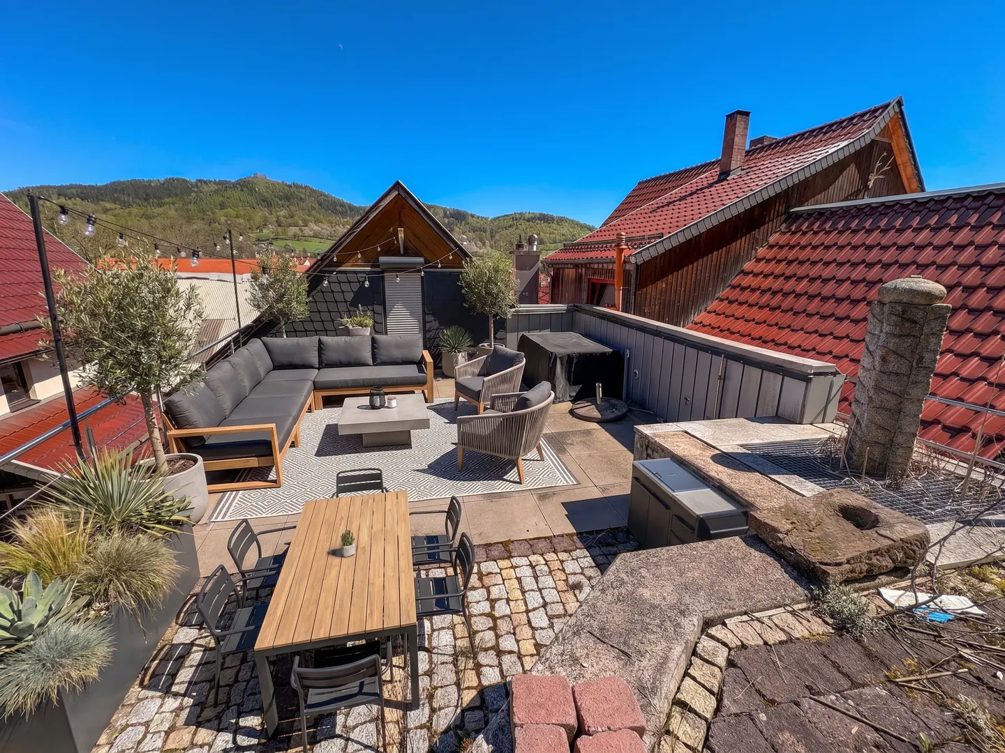 Rooftop patio with gray cushioned seating, a wooden table with chairs, and potted plants, set against a backdrop of red-tiled roofs and green hills.