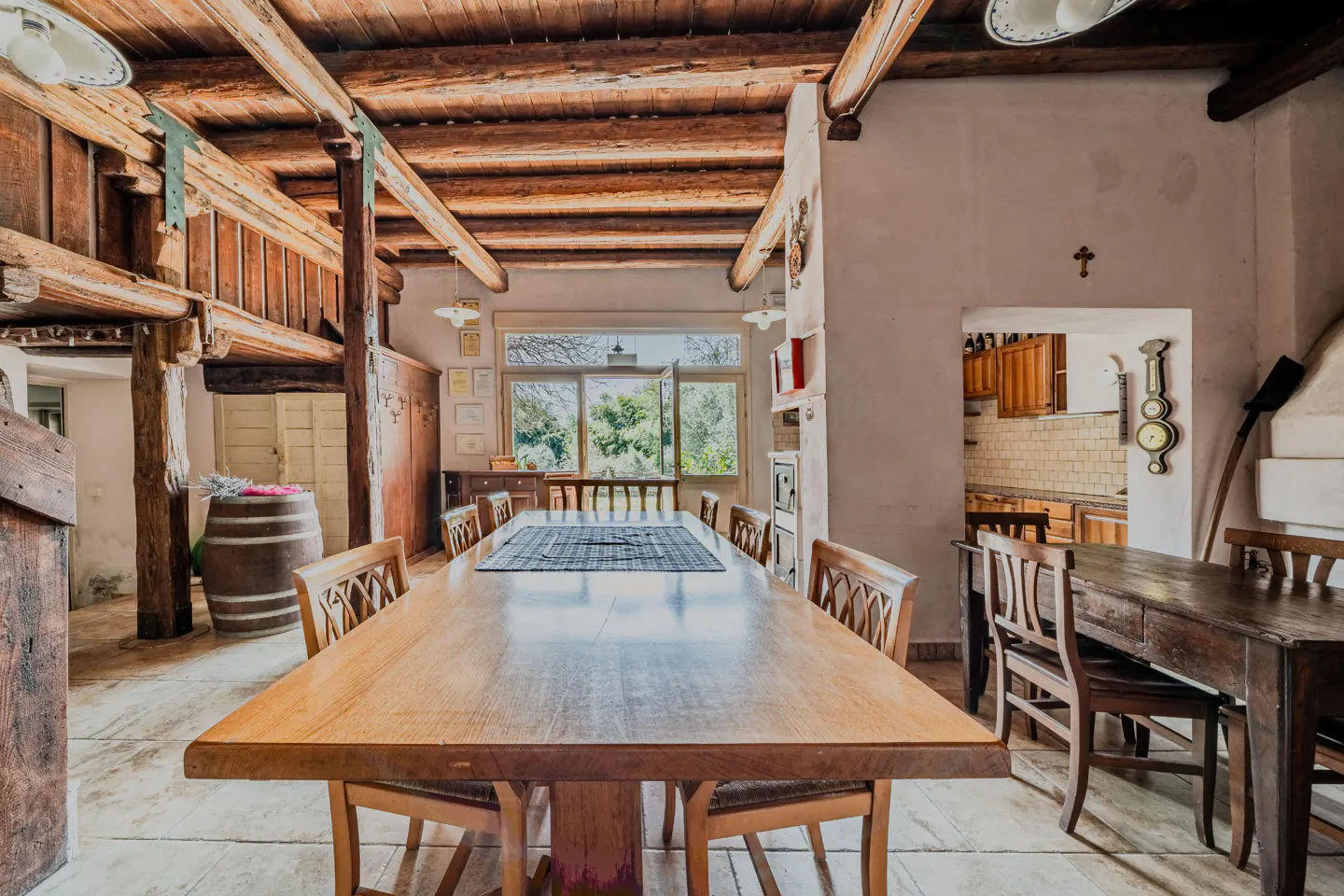 A dining room with a long wooden table and chairs, exposed wood beams, and a view to the outside.