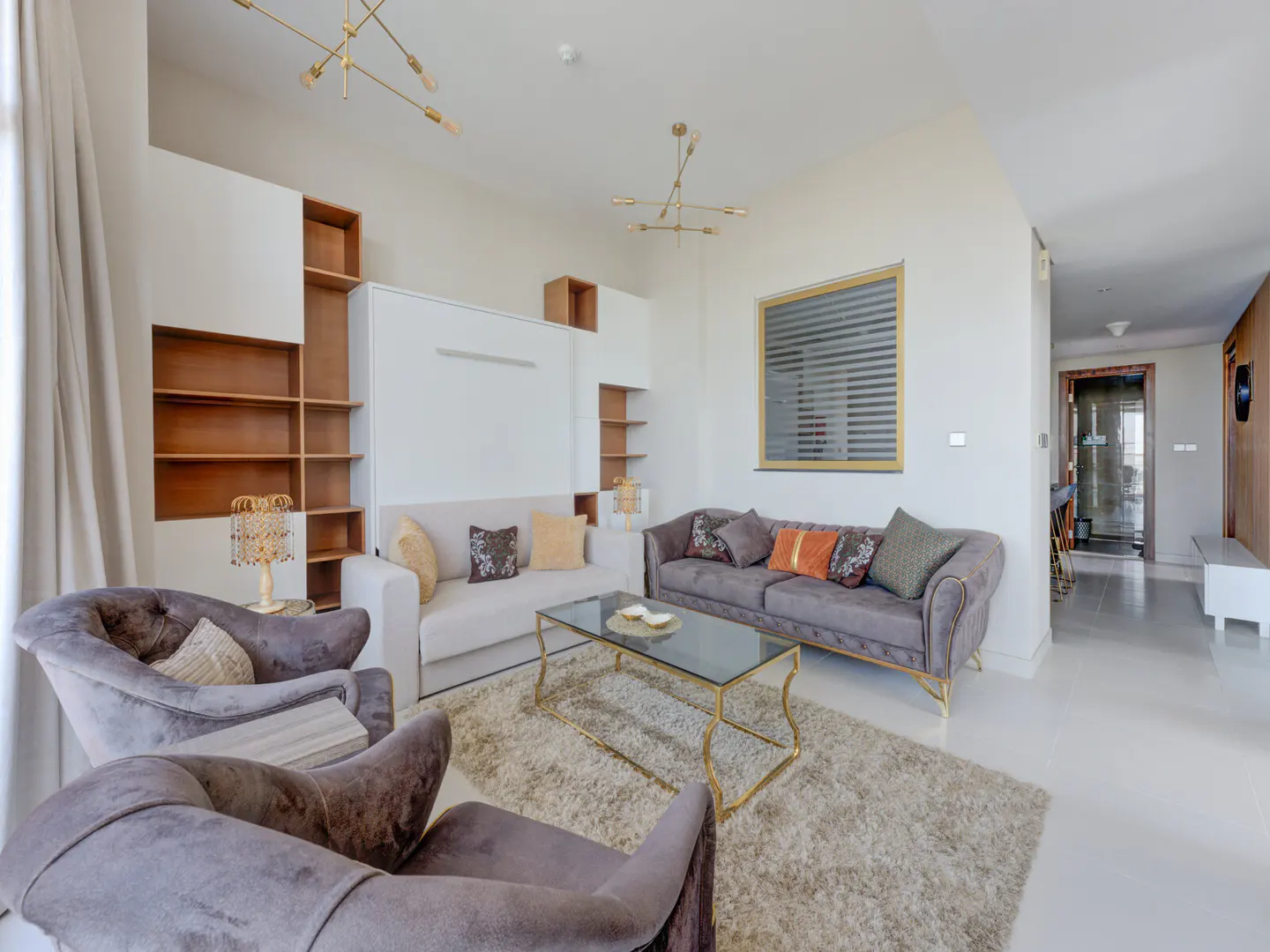Bright living room with two gray armchairs, a white sofa, and a gray sofa. A glass coffee table sits on a shag rug. Built-in shelves and a window are in the background.