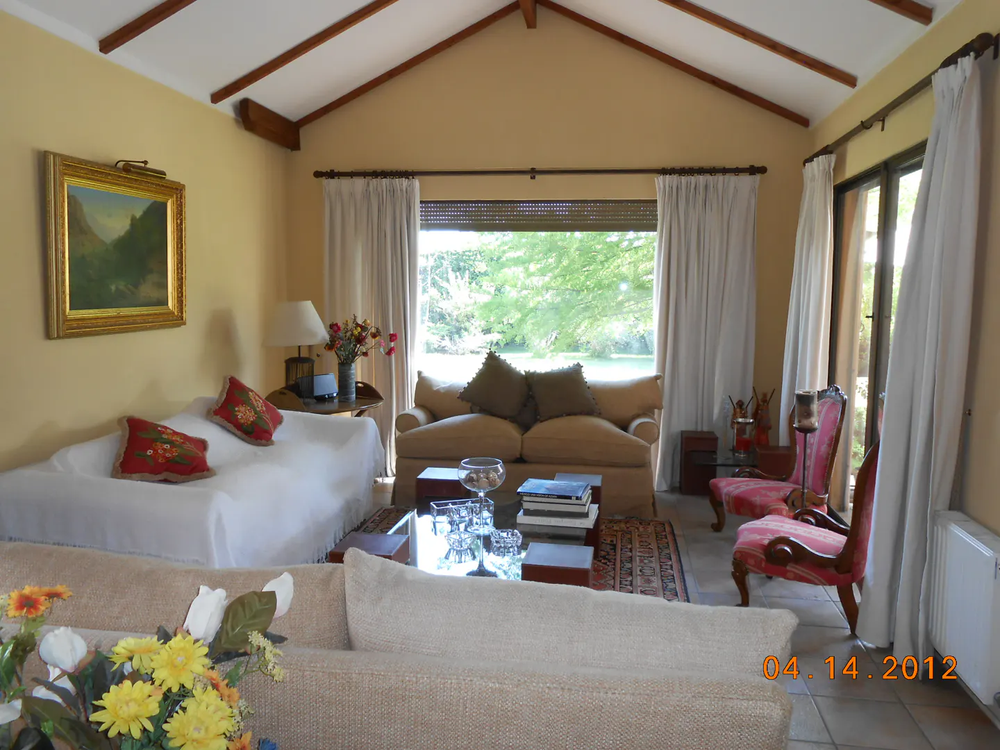 Living room with beige walls, vaulted ceiling, sofas, chairs, and a large window with white curtains overlooking a green landscape.