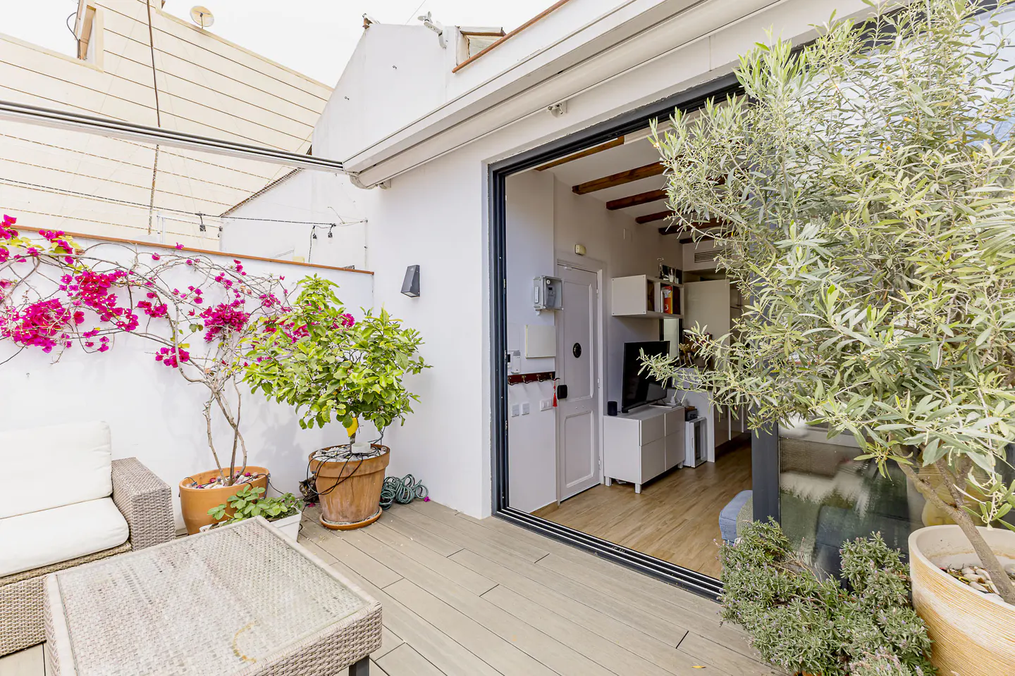 Outdoor patio with sliding glass doors leading to a living room. Potted plants and wicker furniture decorate the patio.