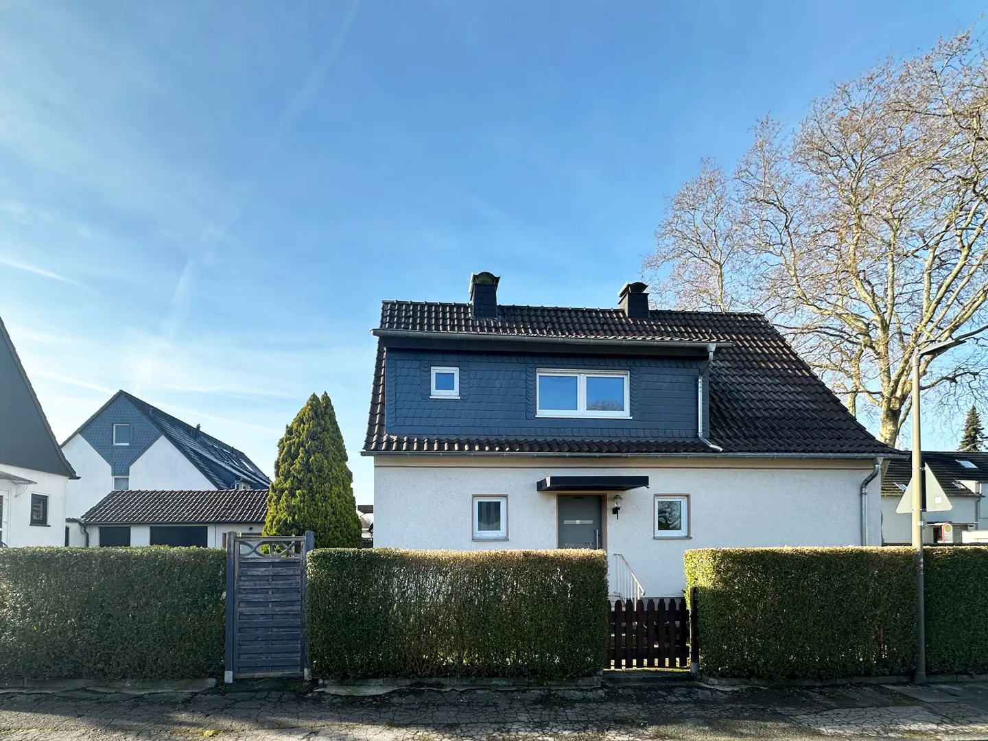 Two-story house with a dark roof and white siding, behind a green hedge and a wooden fence, under a blue sky.