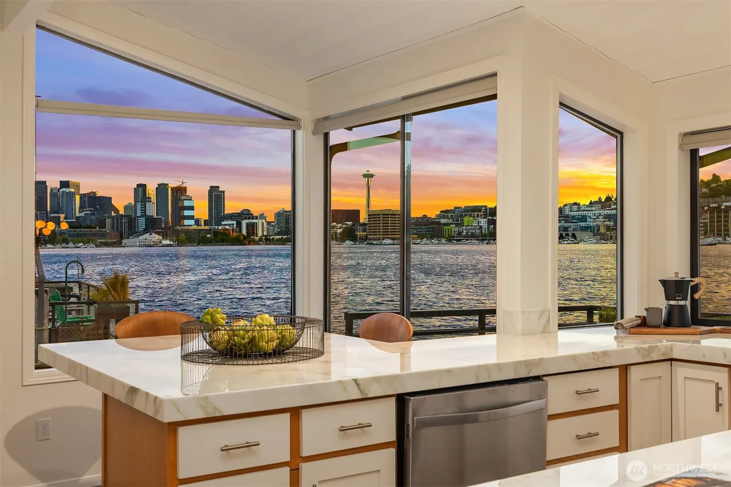 A kitchen island with a marble countertop overlooks the Seattle skyline at sunset through large windows.