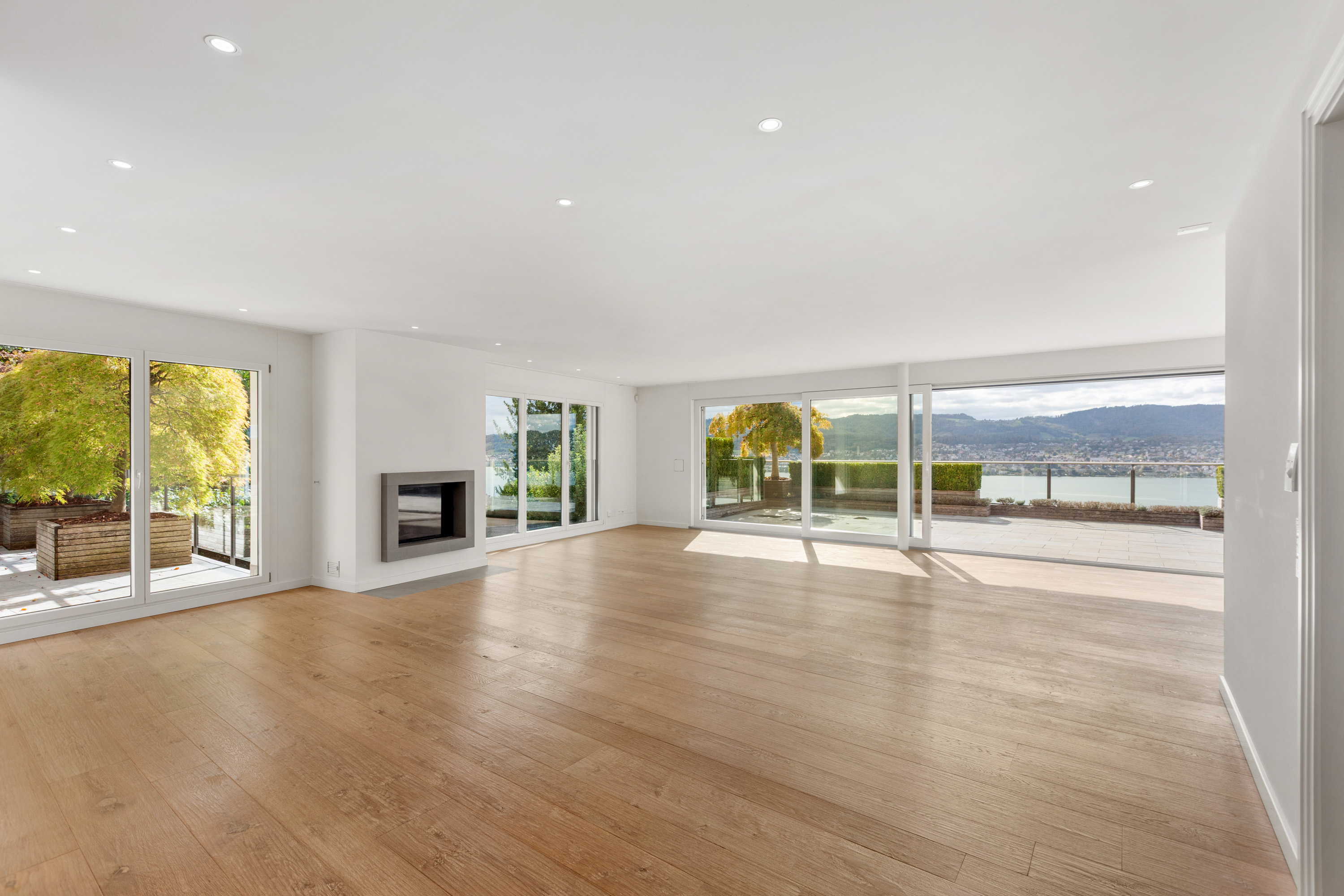 Spacious, minimalist living room with light wood floors, a modern fireplace, and large windows overlooking a lake and hills. [AI-generated text]