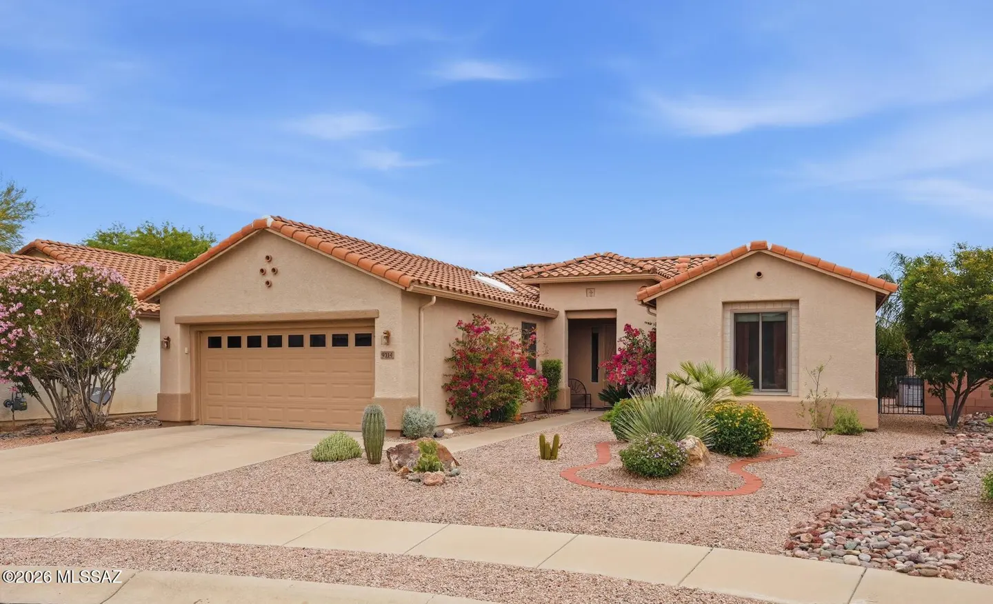 Tan stucco house with a brown garage door and a red tile roof under a blue sky. Desert landscaping with cacti and pink flowers.
