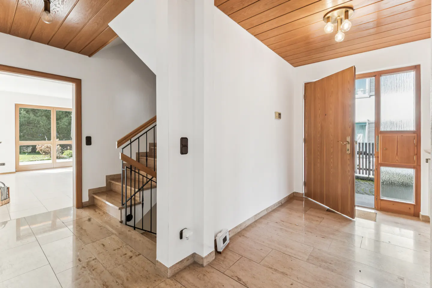 A bright foyer with a wood ceiling, marble floor, and an open front door. A staircase is visible to the left.