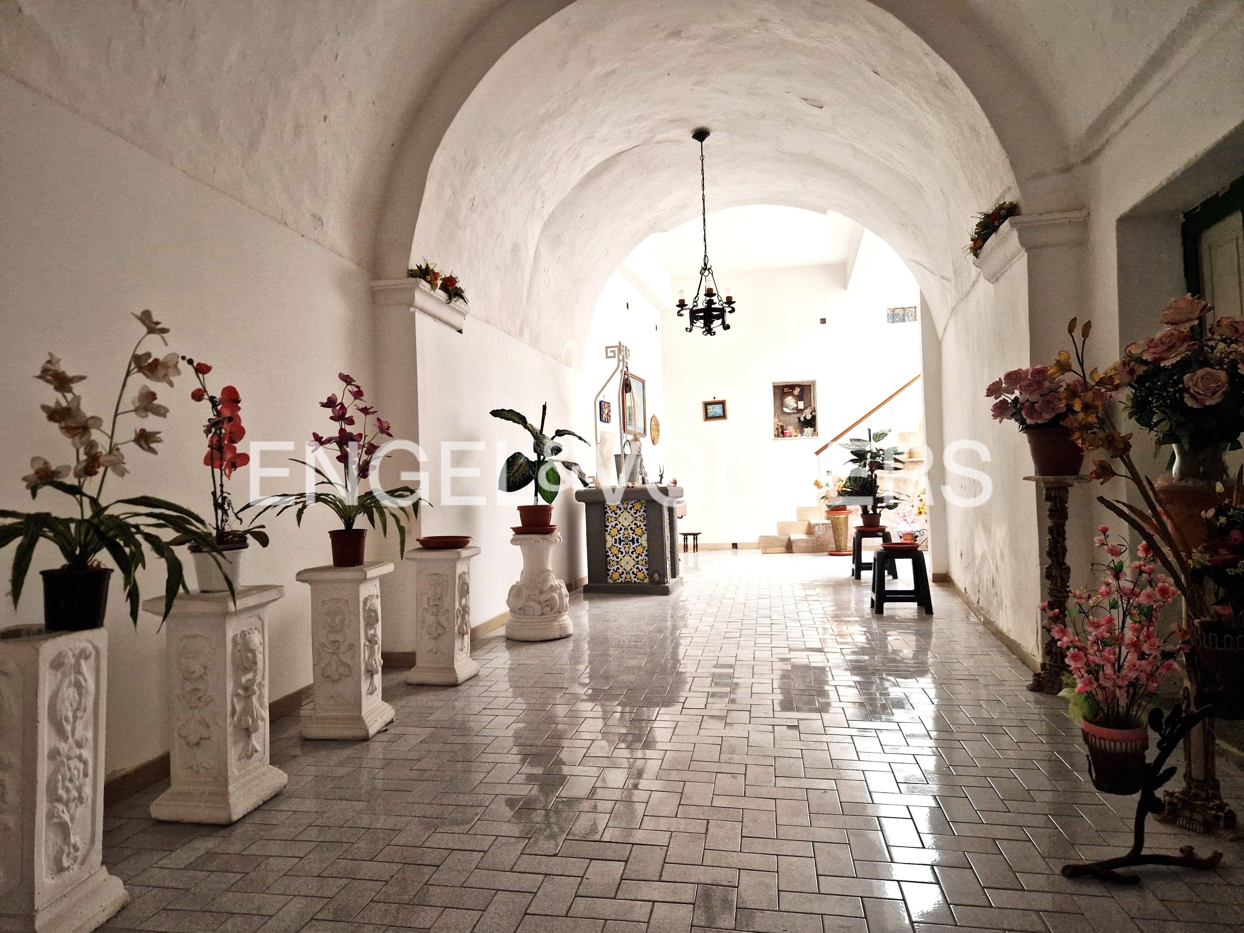 Hallway with arched ceiling, tiled floor, and potted plants on pedestals. A chandelier hangs in the distance.