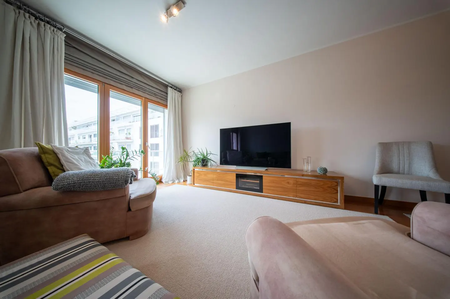 Bright living room with beige carpet, brown sofa, TV on wooden stand, and large windows with white curtains.