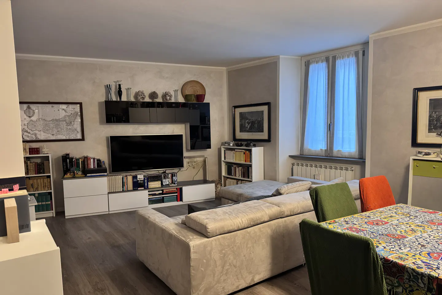 Living room with gray walls, wood floors, a beige sofa, and a table with colorful chairs. A TV and bookshelves are against the wall.